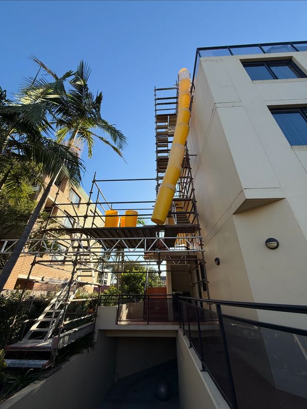 Yellow construction chute attached to scaffolding on a building under clear blue sky.