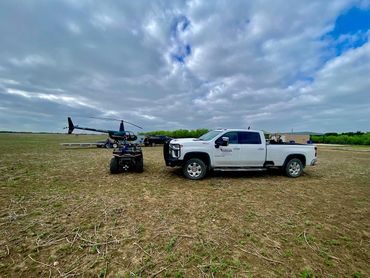 A white pickup truck, ATV, and helicopter parked on a dry field under a cloudy sky.