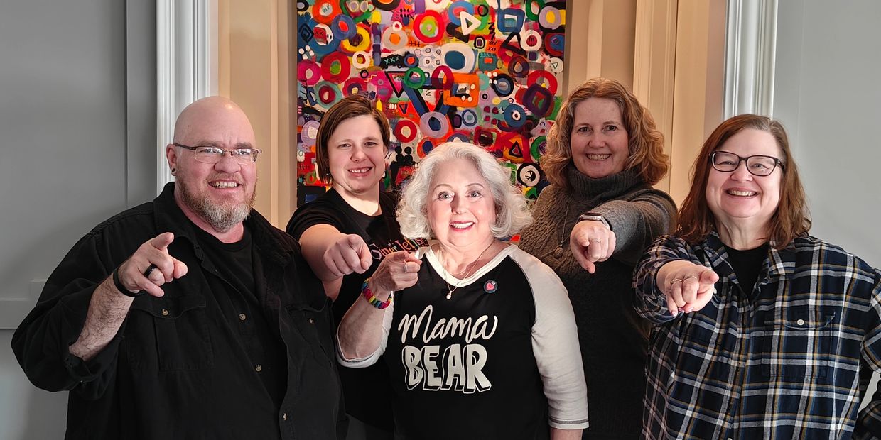 Five smiling people pointing forward, standing in front of colorful abstract art.