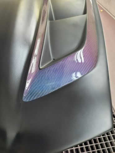 Close-up of a carbon fiber car part with iridescent blue and purple hues.