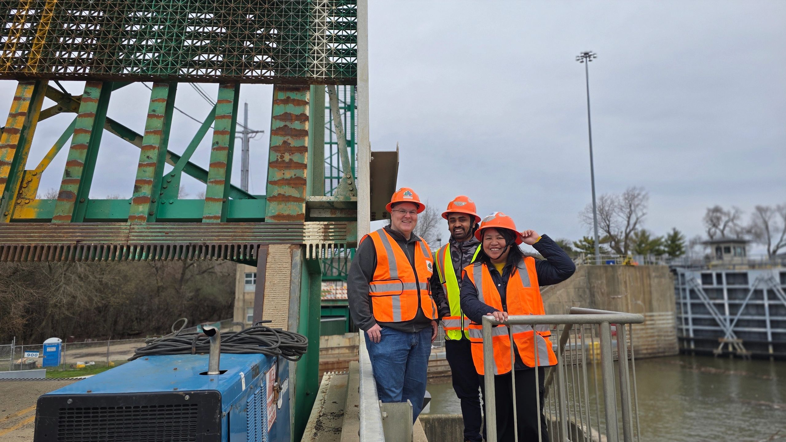 Three construction workers in safety gear smiling at a worksite near a waterway and rusted metal structure.