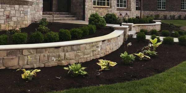 Curved stone retaining wall with plants and fresh mulch in front of a house.