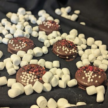 Chocolate-covered cookies decorated with white pearls and surrounded by mini marshmallows on black fabric.