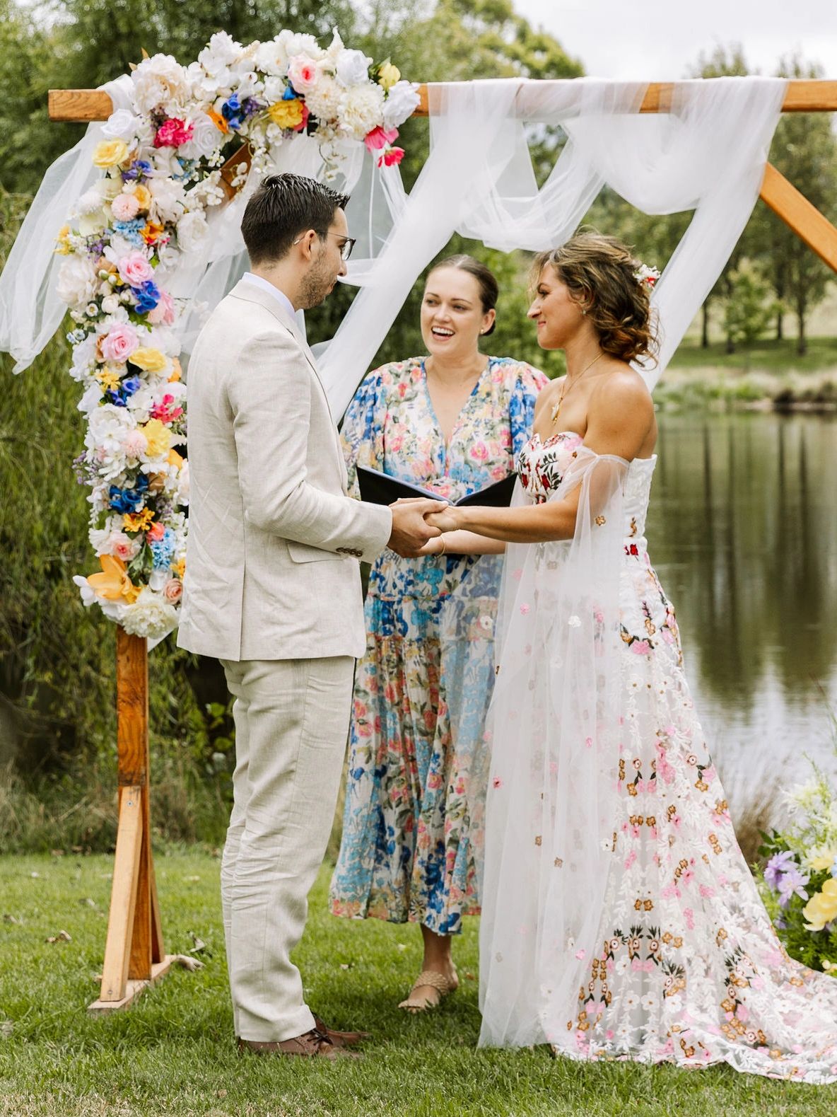 Couple exchanging vows outdoors under a floral arch by a lake.