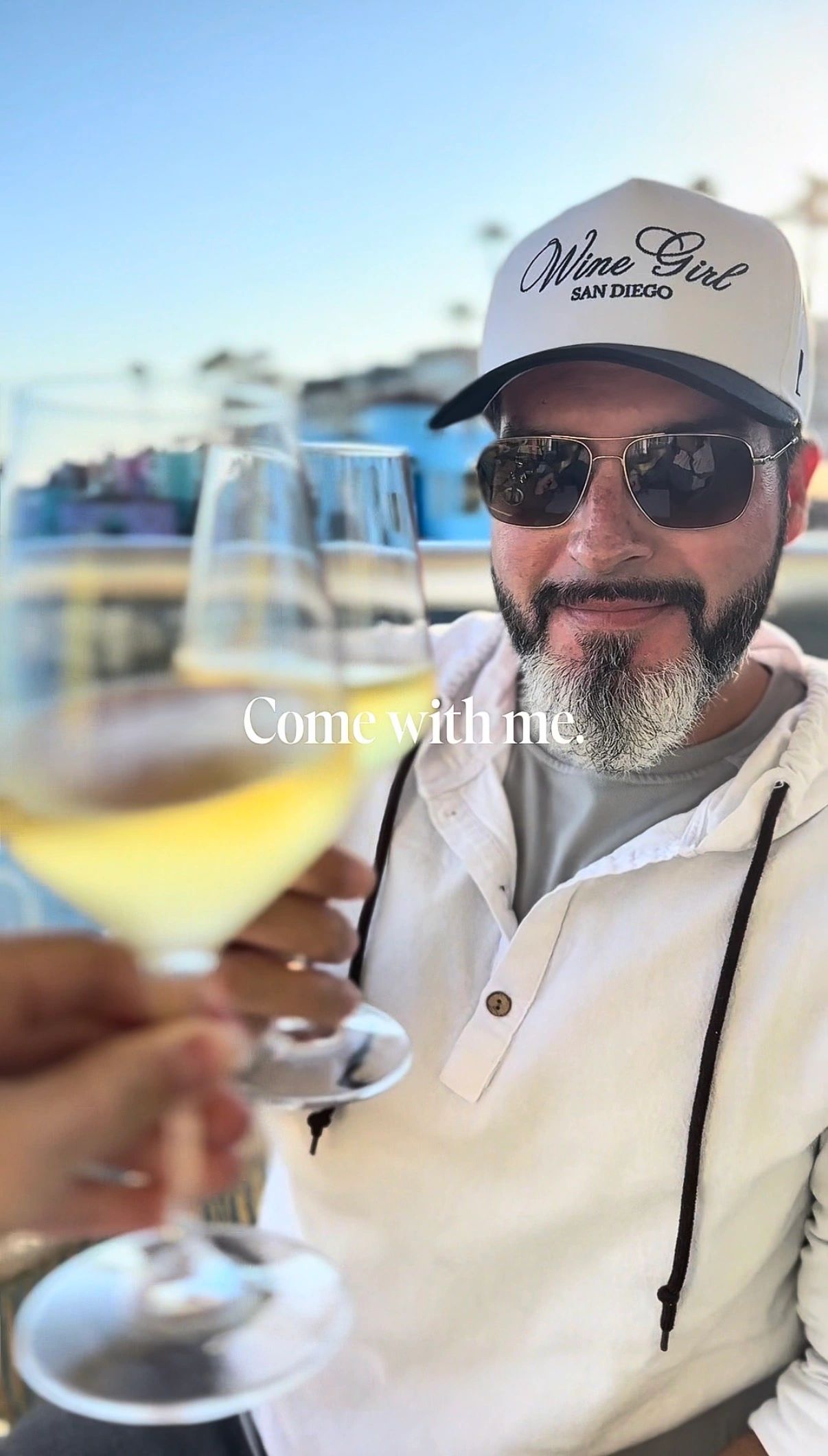 Man toasting with white wine, wearing 'Wine Girl San Diego' cap.