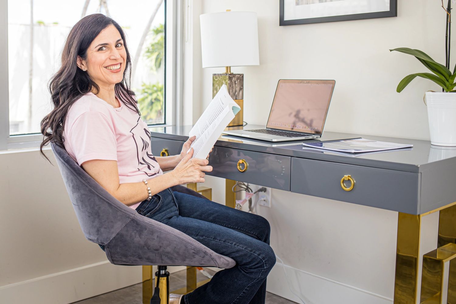 Woman sitting at a desk smiling and holding a book in a bright room.