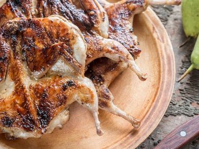 whole flattened roasted quail stacked on a wood serving dish next to a knife and two pairs