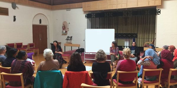 a group of women of different ethnicities sit in a circle listening to two female presenters