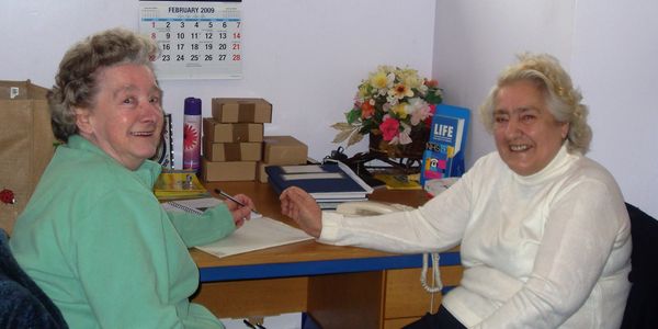 two older white women sit next to a table of paperwork, boxes and flowers. one is filling in a form
