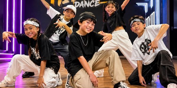 Five kids in streetwear pose confidently in a dance studio with neon lights.
