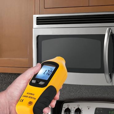 Hand holding a yellow microwave leakage detector near a microwave oven.