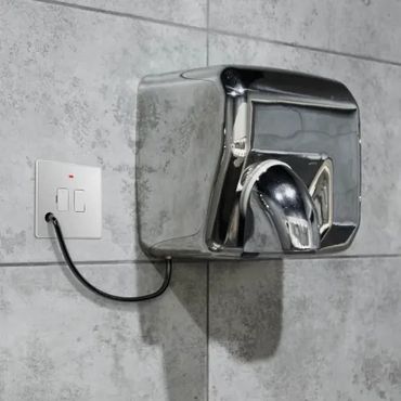 A shiny metal hand dryer mounted on a tiled wall with an electrical cord plugged into a wall socket.