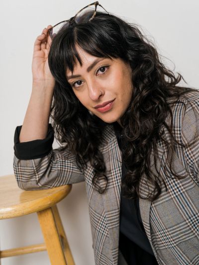Confident woman with curly hair holding glasses, leaning on a wooden stool.