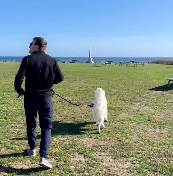 Man walking a white dog on a grassy field near the ocean on a clear day.