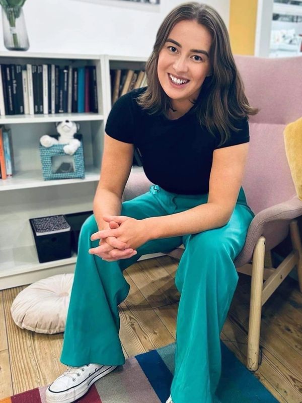 Tegwen, smiling, sitting in a pale pink chair, wearing green pants and black top