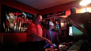 Two men hosting a live show in a Tejano radio studio with microphones and DJ equipment.