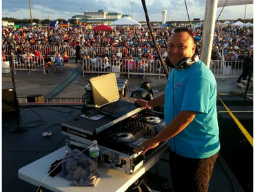 DJ smiling on stage with a large crowd at an outdoor event.
