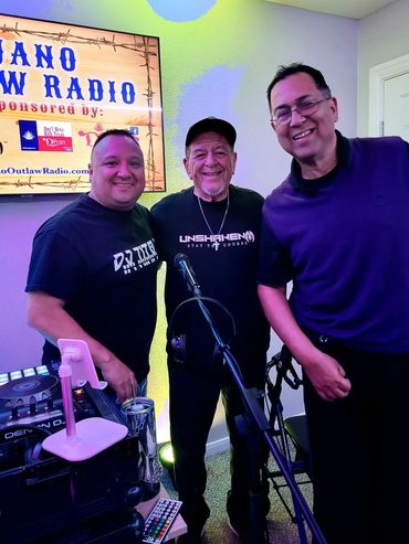 Three men smiling in a radio studio with a microphone and DJ equipment.