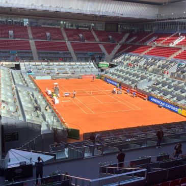 Empty clay tennis court inside a large stadium with some people around.