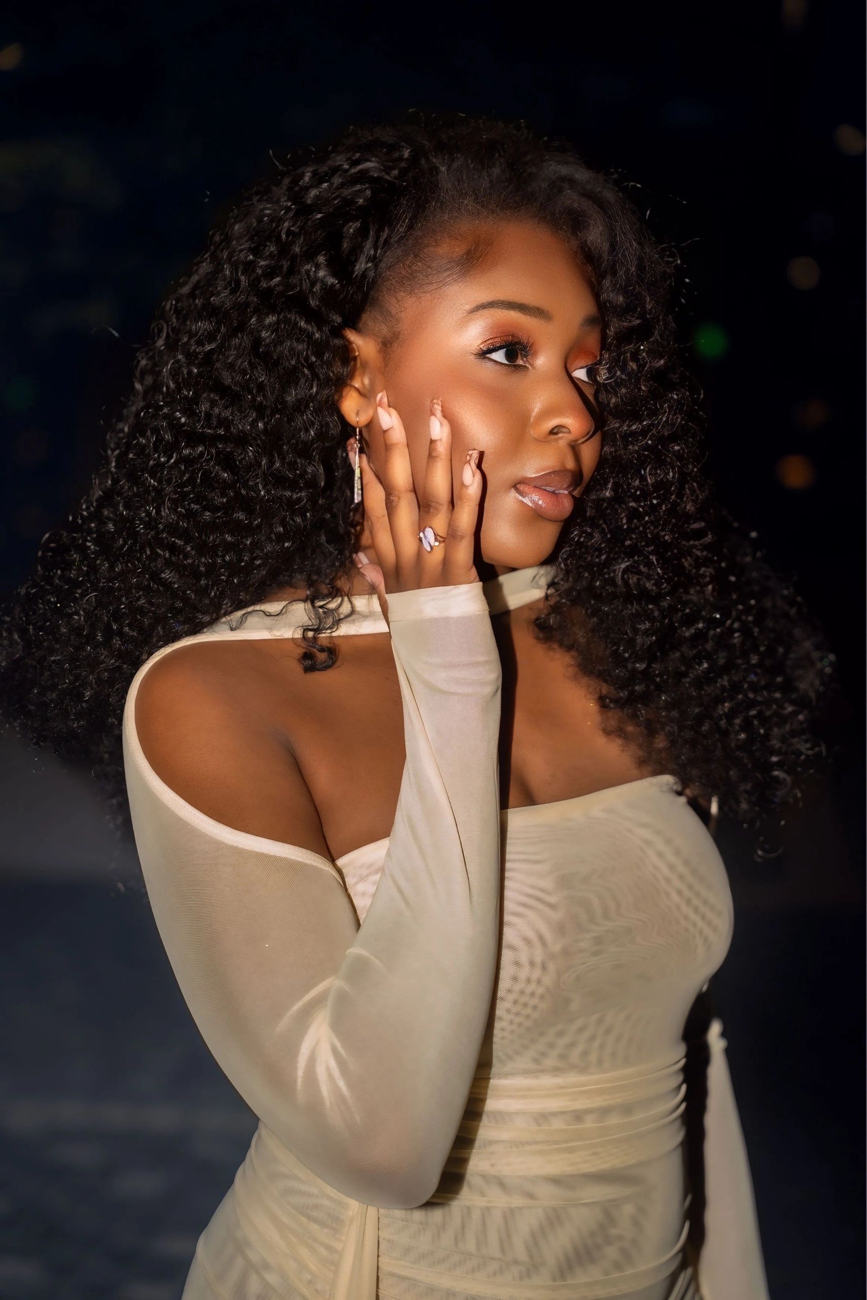 Elegant woman with curly hair in a beige dress, posing thoughtfully with hand on her face.