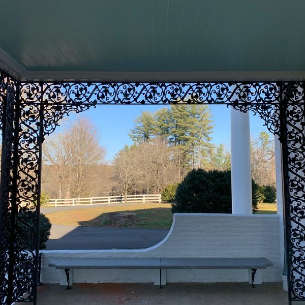Porch with decorative ironwork overlooking trees and a fence.