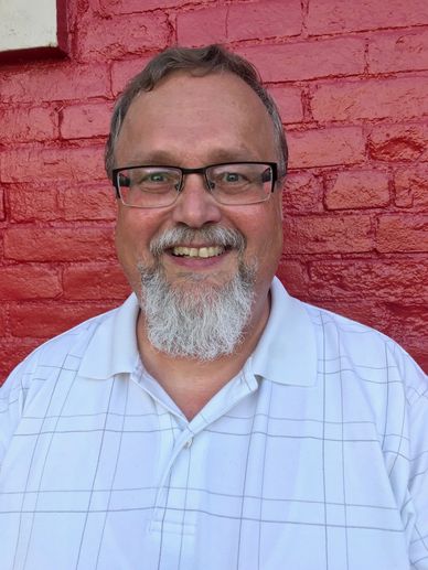 Smiling man with glasses and white beard in front of red brick wall.