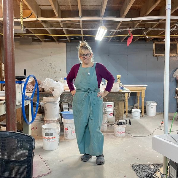 Woman in blue overalls stands in a workshop surrounded by buckets and tools.