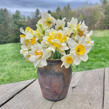 A rustic vase with white and yellow daffodils on a wooden table outdoors.