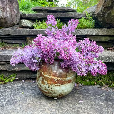 A rustic vase filled with vibrant lilac flowers on stone steps.