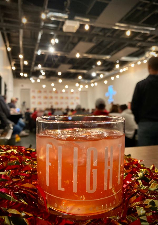 A cocktail glass with "FLIGHT" text filled with a red drink and ice at a lively indoor event.