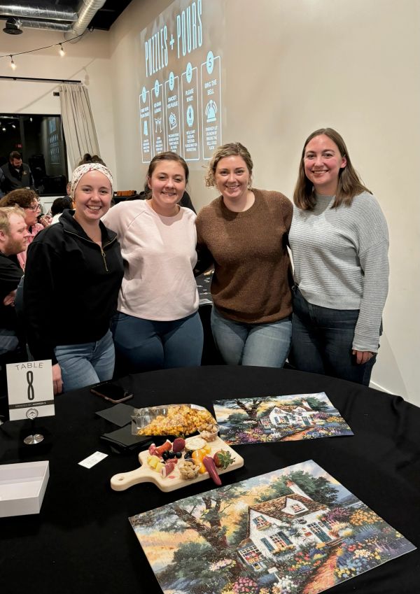 Four women smiling at a table with snacks and artwork at a social gathering.
