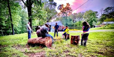 Group of people gathered around a campfire in a green outdoor setting at sunset.