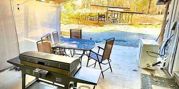 Covered patio with grill, table, and chairs overlooking a wooded backyard.