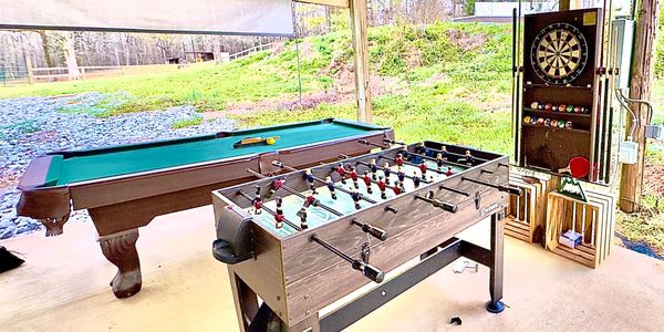 Outdoor game area with foosball, pool table, and dartboard under a wooden shelter.