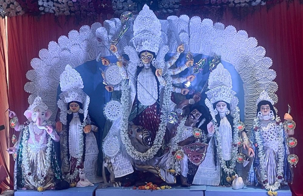 Durga Puja pandal in Kolkata decorated with elaborate lights and artwork, featuring a majestic idol of Goddess Durga slaying Mahishasura, surrounded by Saraswati, Lakshmi, Kartikeya, and Ganesha. Crowds of devotees dressed in festive attire gather for evening prayers, with drums (dhak) playing and incense smoke filling the air.