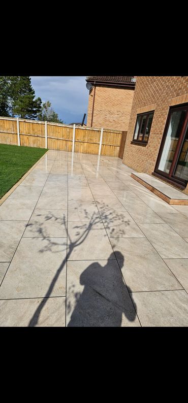 Shadow of a person holding a branch on a tiled patio next to a brick house and wooden fence.