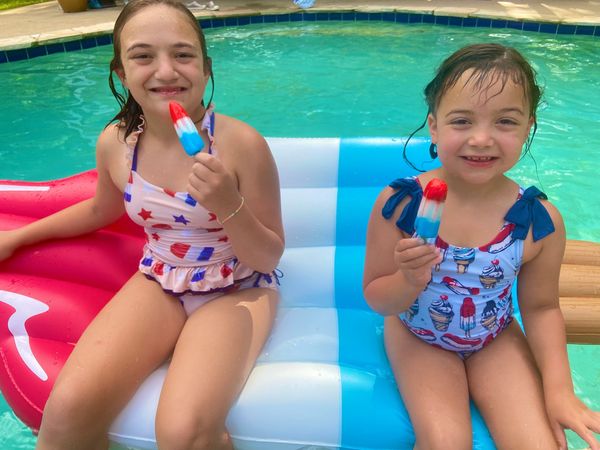 Two girls in swimsuits holding red, white, and blue popsicles by the pool.