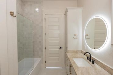 Modern bathroom with beige tones, a round illuminated mirror, and a glass-enclosed bathtub.