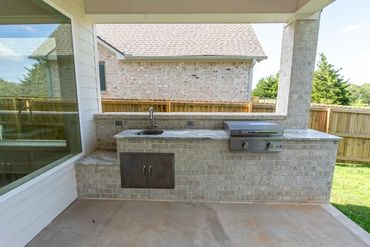 Outdoor kitchen with brick countertop, sink, and grill under a covered patio.