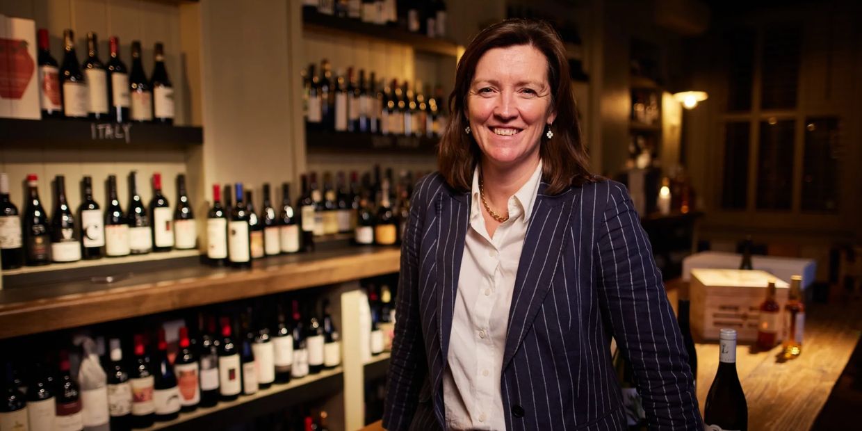 Woman smiling in a wine shop with shelves of bottles behind her.