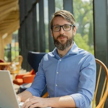 Man with glasses working on laptop in a cozy, sunlit cafe.
