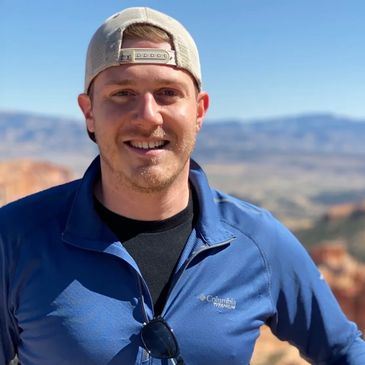 Smiling man in a blue jacket and beige cap outdoors with sunglasses hanging from his jacket.