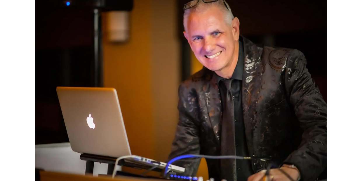 DJ in a black jacket & shirt plays on a macbook pro, surrounded by dancers at a reception.