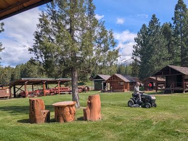 man riding lawn mower, two log chairs and table in foreground, museum building and exhibits in backg