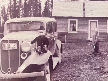 young child sitting on a parked car in front of log building