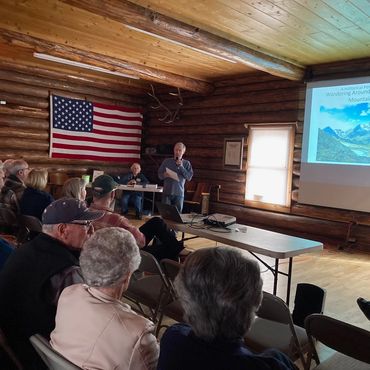 Man standing in front of flag and projection screen, holding microphone
