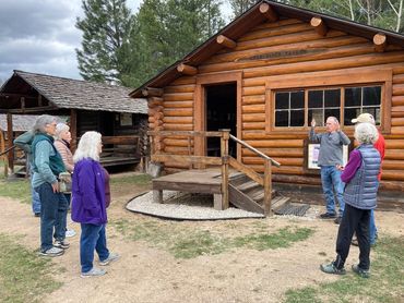 man speaking to group, outside historic log cabin