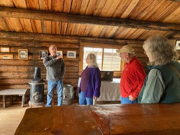 man speaking to three people, inside historic log cabin