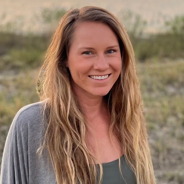 Smiling woman with long blonde hair standing outdoors at sunset by a lake.