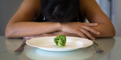 Person with head down in front of a plate with a single broccoli floret.